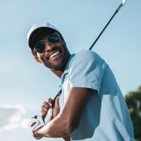 Smiling african american man in cap and sunglasses holding club and playing golf