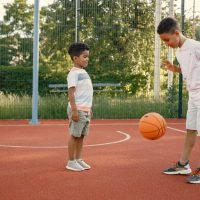 Two multiracional brothers playing basketball on a court near the park. Boys wearing white t-shirts. Older brother teach little one how to play basketball.